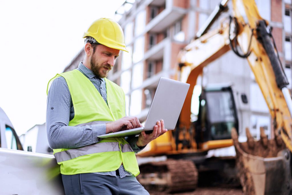 A commercial building official reviews certificate of occupancy requirements on a laptop at a multifamily jobsite featuring an excavator.