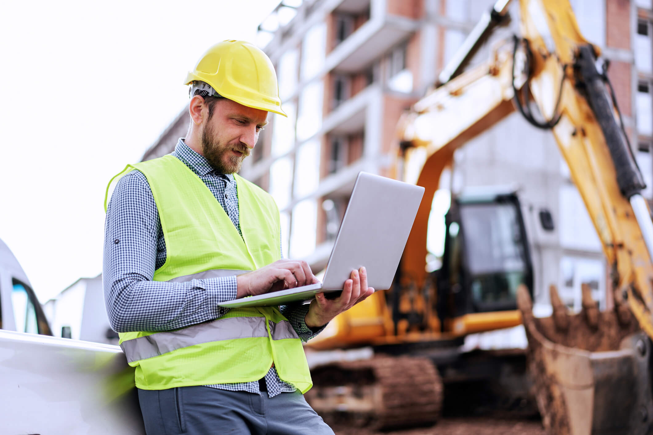 A commercial building official reviews certificate of occupancy requirements on a laptop at a multifamily jobsite featuring an excavator.