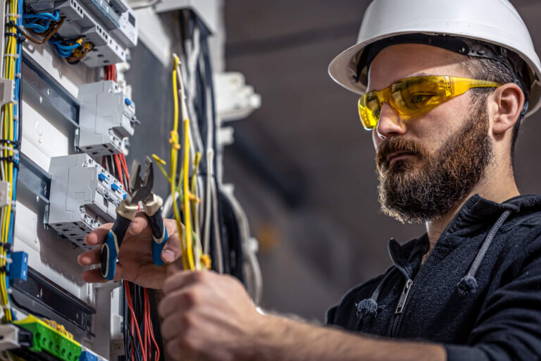 Licensed electrician in hard hat and safety glasses inspecting an electrical service panel with pliers
