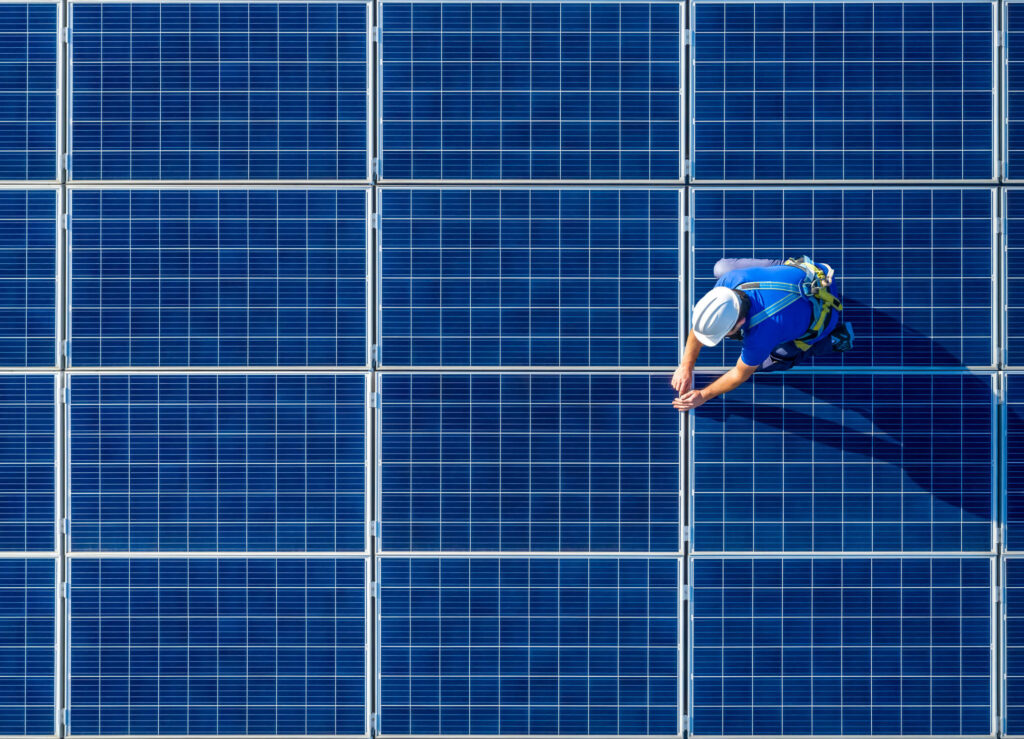 Solar panel technician in safety harness inspecting photovoltaic panels on a rooftop solar installation.
