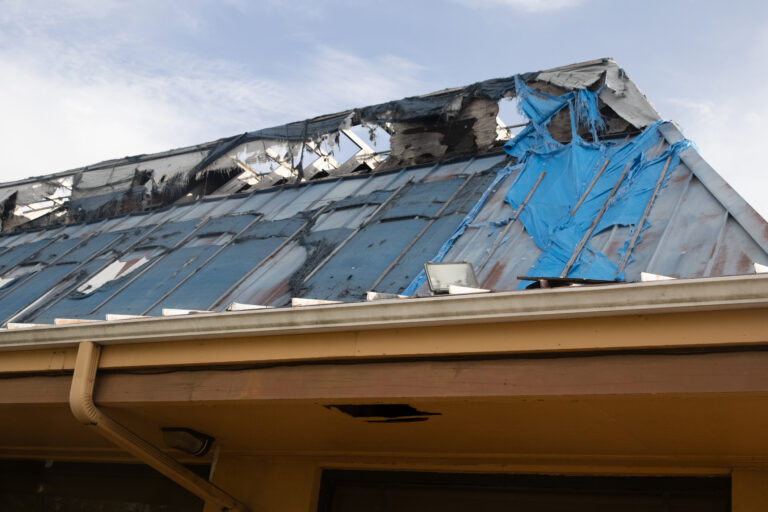 Storm-damaged metal roof with torn panels and blue tarp covering wind damage on a residential structure